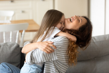 Close up happy attractive mother laughing hugging cute preschool daughter. Smiling young mom closed eyes embracing little girl playing in free time together sitting on couch at home.