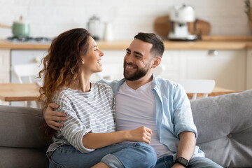 Happy young couple of man and woman embracing look at each other sitting on couch in living room. Attractive smiling husband and wife laughing having fun free time together. - Powered by Adobe