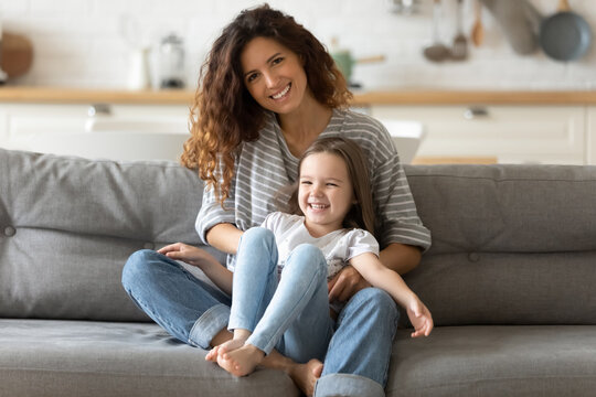 Close up portrait picture of smiling young mother with daughter sitting on couch at home. Happy parent with little child hugging looking at camera posing for photo in living room.