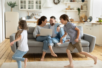 Happy young couple sitting at couch near running children using laptop in modern living room....
