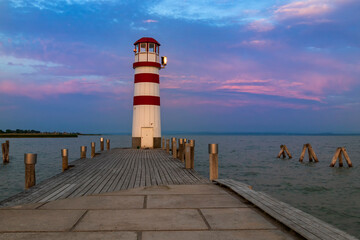 A pier leading to the lake and a red and white lighthouse on Lake Neusiedl in Podersdorf, Austria. In the background is a dramatic sky at sunrise.