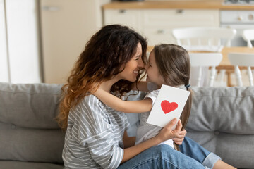 Close up young mother holding gift postcard with heart embracing little daughter congratulate young diverse mom with mothers day or birthday. Adorable child making surprise to mum at home.