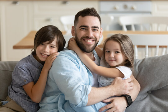 Close up headshot portrait picture of happy young father with children sitting on couch at home. Smiling parents with little kids hugging looking at camera posing for photo in living room.