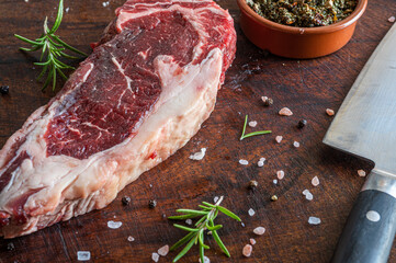 Close up of raw vaca paraguaya steak with rosemary, himalayan salt, black pepper grains and a knife over a wooden board
