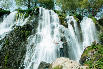 A man with his hands open stands by a waterfall