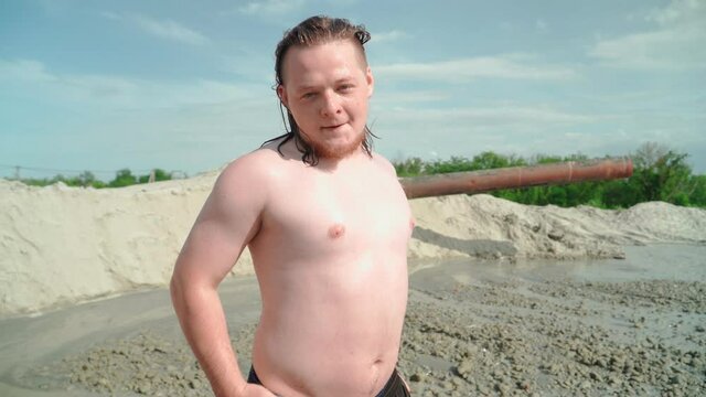 Funny Fat Wet Man with Long Hair Posing. Against the background flows dirty water and piles of sand