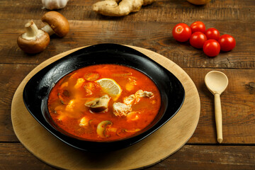 Traditional thai tom yam soup with chicken in a black plate on a wooden stand with a spoon and vegetables nearby.