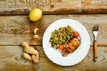 On a wooden table is a white plate with baked trout steak and a side dish of quinoa with vegetables and a fork next to it.