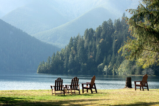 Adirondack chairs in the grass on the side of a lake overlooking pine trees and mountains