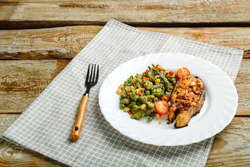 On a linen napkin is a white plate with baked trout steak and a side dish of quinoa with vegetables and a fork next to it.