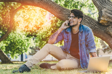 Young handsome freelancer working on a laptop and talking to the phone outdoors. Outdoor work concept.