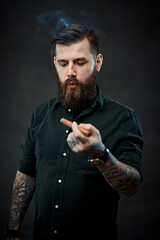 Stylish bearded man smokes a cigar. Studio portrait of a handsome tattooed hipster guy who smokes a cigar