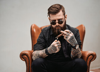 Cool tattooed bearded man lights a cigar while sitting on a vintage chair in studio