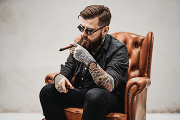 Bearded tattooed man with stylish haircut in sunglasses who smokes a cigar while sitting on a...