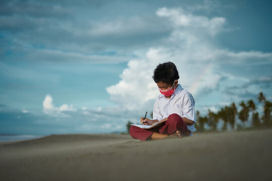 Schoolboy wearing school uniform and face masks writing and studying at the beach. homeschooling during a virus outbreak - Powered by Adobe