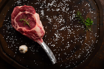 Top view raw tomahawk beef steak with ingredients for grilling with a spice rub, fresh rosemary and salt , on a table with dark wood texture and space for writing