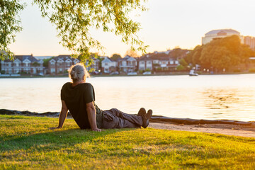 Back view gray hair mature man sitting on the green grass at the bank of park's lake and enjoying...