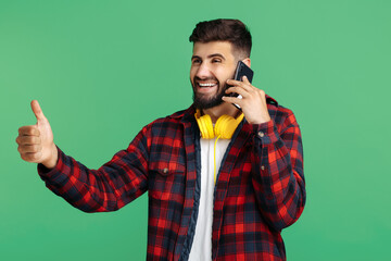 Trendy bearded hipster young man talk on smartphone and show thumb up over green background.