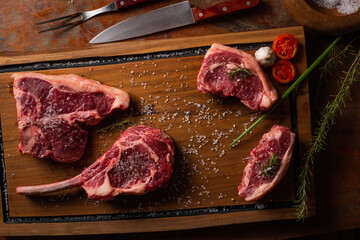 Top view raw tomahawk, loin,  t-bone beef steak with ingredients for grilling with a spice rub, fresh rosemary and salt , on a table with dark wood texture and space for writing.