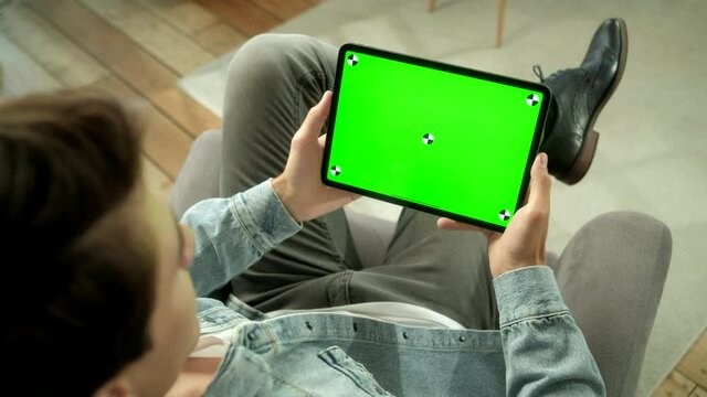 View From the Shoulder of Man Holding and Using Hand Gestures on Green Mock-up Screen Digital Tablet Computer Sitting on a Chair. Male Buying stuff or Browsing. Tapping on Center Screen