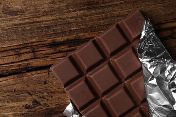 Tasty milk chocolate bar wrapped in foil on wooden table, top view