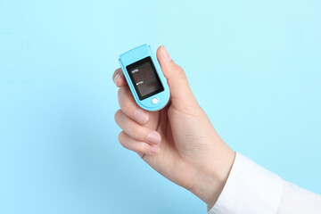 Woman holding modern fingertip pulse oximeter on light blue background, closeup
