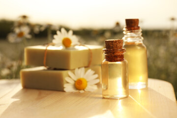 Fototapeta na wymiar Bottles of chamomile essential oil and soap bars on wooden table in field