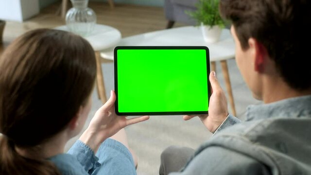 Over The Shoulder View Young Couple at Home Sitting on a Sofa Using With Green Mock-up Screen Tablet. Man and Woman Using Touchscreen Device, Browsing Internet. Concept of Green Screen and Chroma Key.