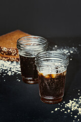 two steaming mug with dripping drops, full of dark carbonated drink with lush foam on black background with scattered wheat flakes and black rye bread on background. Traditional drink kvass