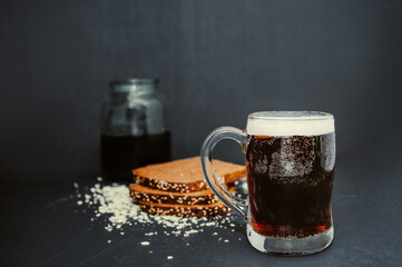 beautiful tall steaming mug with dripping drops, full of dark carbonated drink with lush foam on black background with scattered wheat flakes and black rye bread on background. Traditional drink kvass