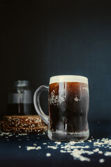 beautiful tall steaming mug with dripping drops, full of dark carbonated drink with lush foam on black background with scattered wheat flakes and black rye bread on background. Traditional drink kvass