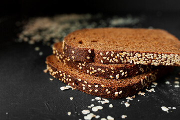 Three slices of black rye whole grain bread with sesame seeds on a black background with scattered wheat flakes