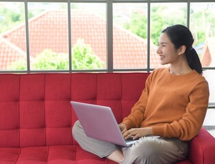 Naklejka premium Asia woman is smiling with working to laptop and sitting on red sofa at her room