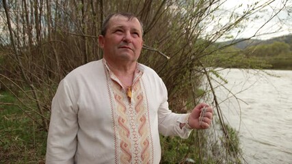 Old grandfather senior elderly man standing on the river bank in an embroidered shirt is conceived, remembers, recalling past and kisses silver cross with a chain