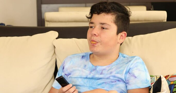 Teenage boy with tv remote controller chewing bubble gum and watching tv at home