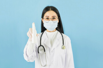 Smart young asian female doctor in lab coat with Medical face mask,white latex medical gloves and stethoscope,Holding a bottle of spray alcohol against blue background,health care concept