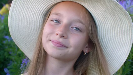 Beautiful happy teenage girl in hat smiling at camera outdoors. Close-up portrait of adorable young girl resting on green meadow with blooming flowers at summertime