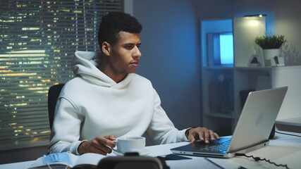 Handsome multiracial man drinking a cup of coffee and working on computer at night. There are skyscrapers in the background.