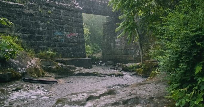 4K Rain Falling on Natural Forest Stream Under Stone Railroad Bridge, Daytime Storm on Wooded River