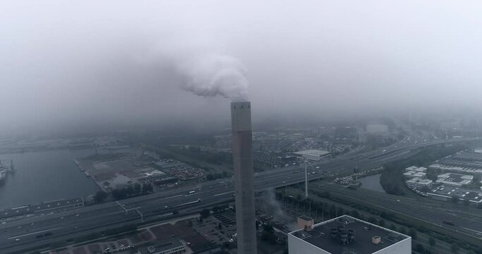Backward aerial reveal of smoking chimney of coal fired plant in industrial port with highway on background Amsterdam Netherlands