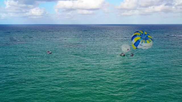 Two people parasailing together in yellow and blue parachute in open green ocean waters, circle aerial