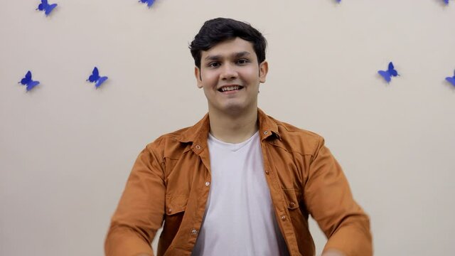 Attractive young teenager showing thumbs-up signs while smiling at the camera. Portrait of a handsome Indian guy happily making ok gestures while sitting against a decorated wall - expressions concept