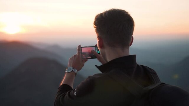 Slow motion shot of a young man taking a picture with his smartphone from the top of a mountain