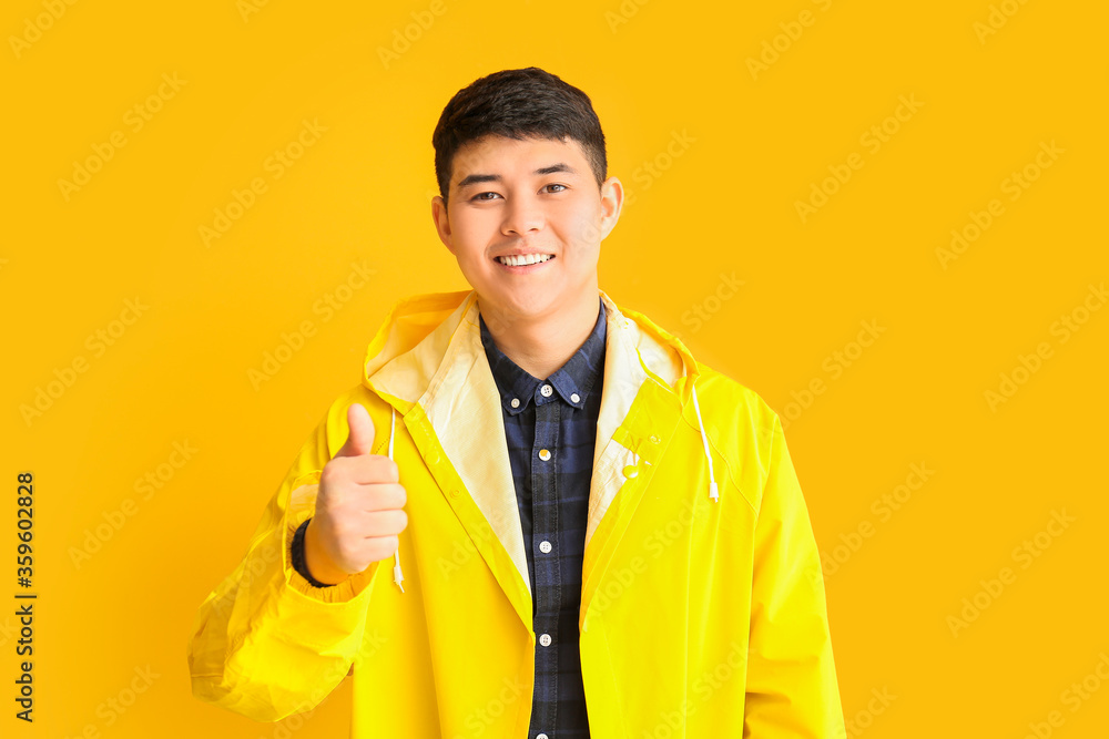 Wall mural Young Asian man in raincoat showing thumb-up on color background