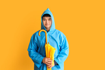 Young Asian man in raincoat and with umbrella on color background