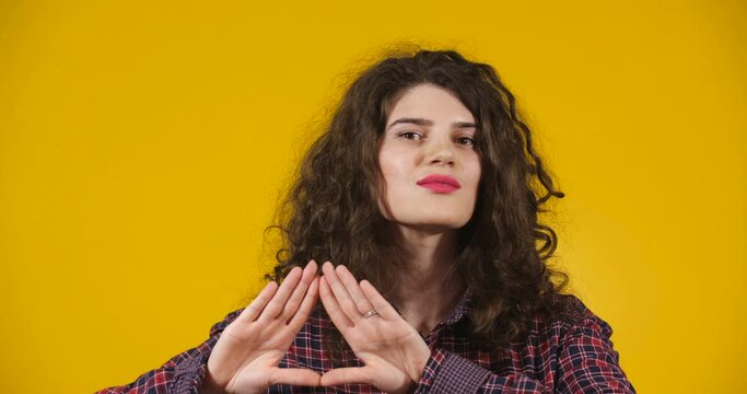 Close-up attractive plus-size model posing. A curly-haired brunette on a bright yellow studio background demonstrating different emotions. Girl making a delta sign with her hands