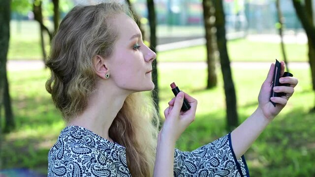 Video caucasian blonde girl paints lips with lipstick in the park. Model posing in clear weather.