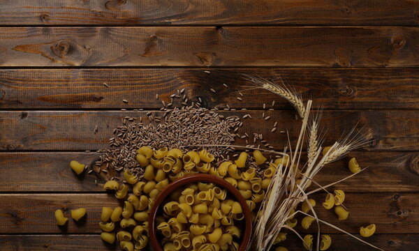Wheat ears, spelt grains, kernels and macaroni lumaconi rigati with clay bowl, pot on wooden plank table background, top view