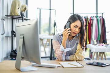 Young Asian woman sitting in front of computer talking on a phone call in a clothing shop, looking stressful and unhappy.
