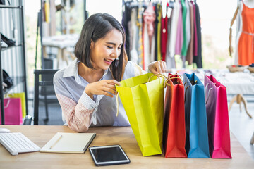 Young Asian girl smiling sitting at a desk inside a clothing store with a notebook and tablet, looking inside multicolor shopping bags.